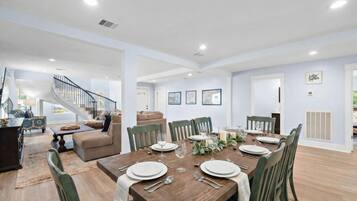 A spacious dining area with a wooden table set for six, surrounded by green chairs. A staircase is visible in the background leading to an upper level.