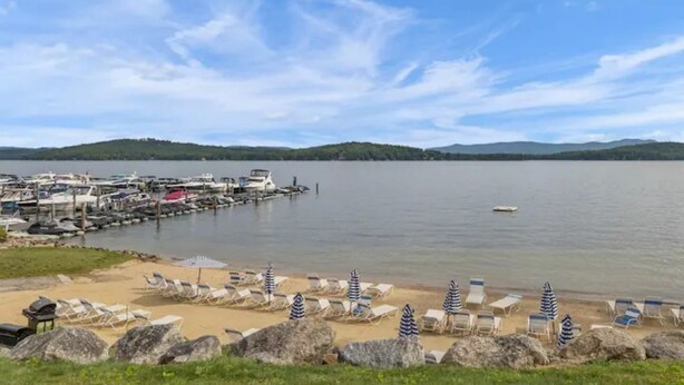 A marina with boats docked, a sandy beach with lounge chairs and umbrellas, and a calm lake with hills in the background. - Cozy Winter Escape (Laconia)