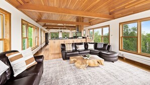 A spacious living area with a large sectional sofa, a wooden coffee table, and a view of the kitchen and outdoor greenery through large windows.