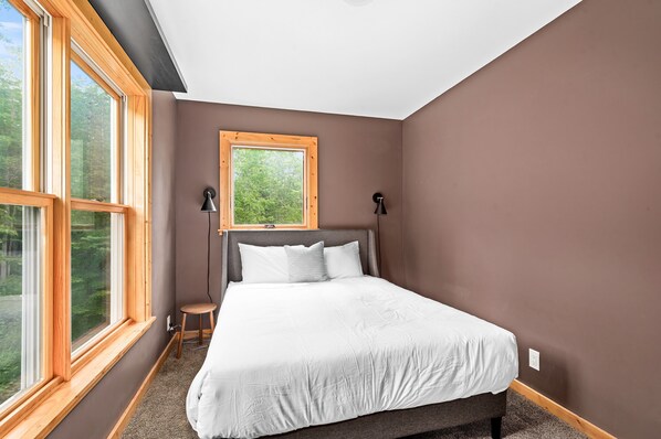 A bedroom with a large window, a bed with white bedding, a small stool, and two wall-mounted lamps.
