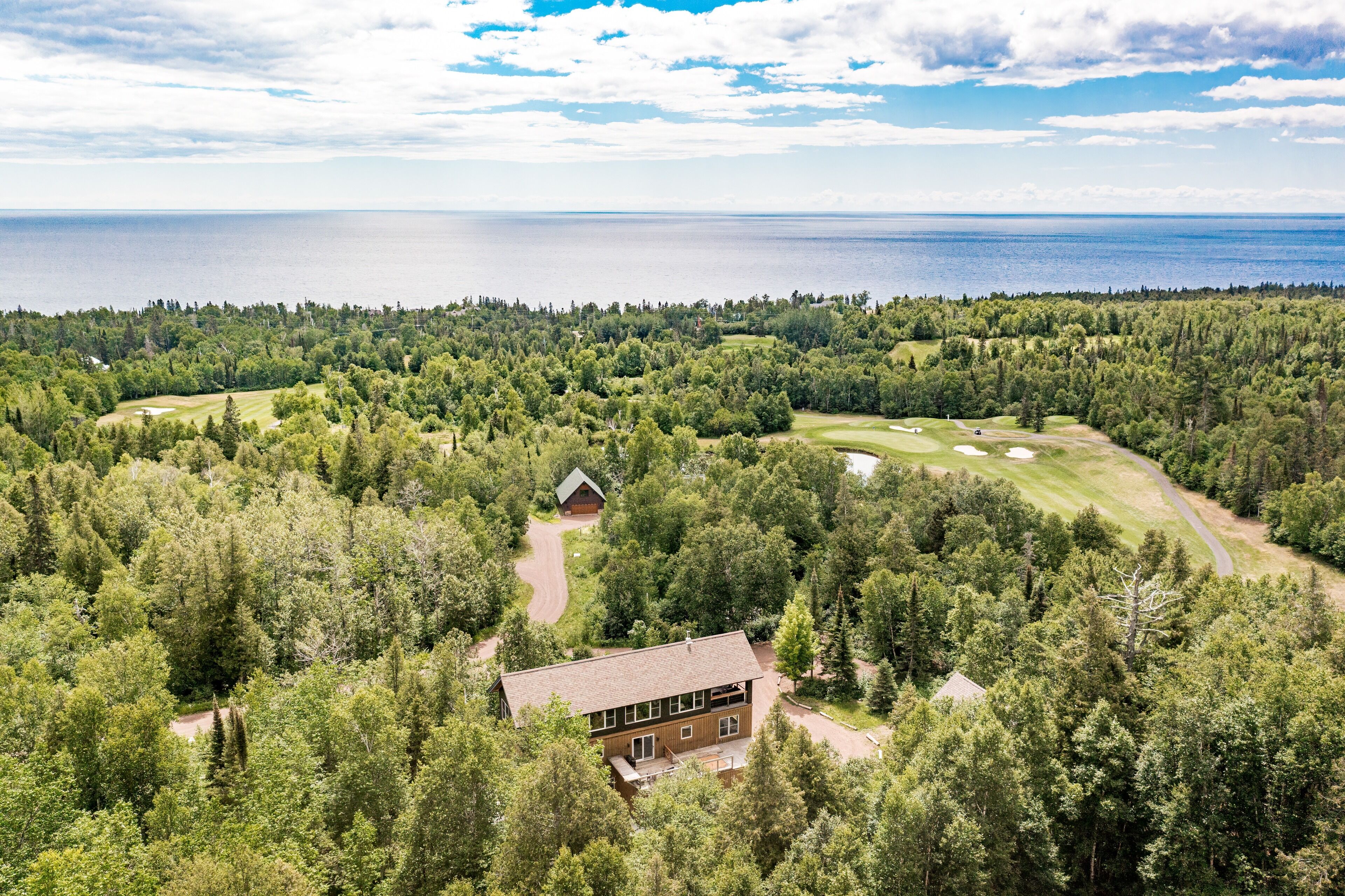Vue aérienne d’une propriété comprenant un grand bâtiment entouré d’une forêt dense, un terrain de golf et une étendue d’eau en arrière-plan.