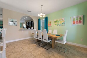 A dining area with a wooden table, white chairs, and a chandelier. There are framed artworks on the walls and a kitchen in the background. - Orange Pineapple at Solterra by Shine Villas 411 (Davenport)