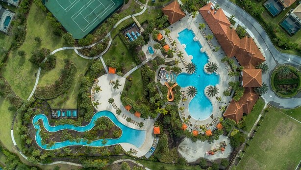 An aerial view of a resort with a large central pool, a tennis court, and a winding pool.