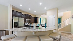 A kitchen with dark cabinets, a stainless steel refrigerator, a microwave, and a granite countertop. There are four bar stools and a trash can.
