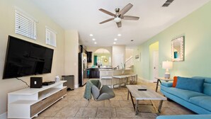 A living room with a flat-screen TV, a ceiling fan, a blue sofa, a coffee table, and a kitchen area in the background.