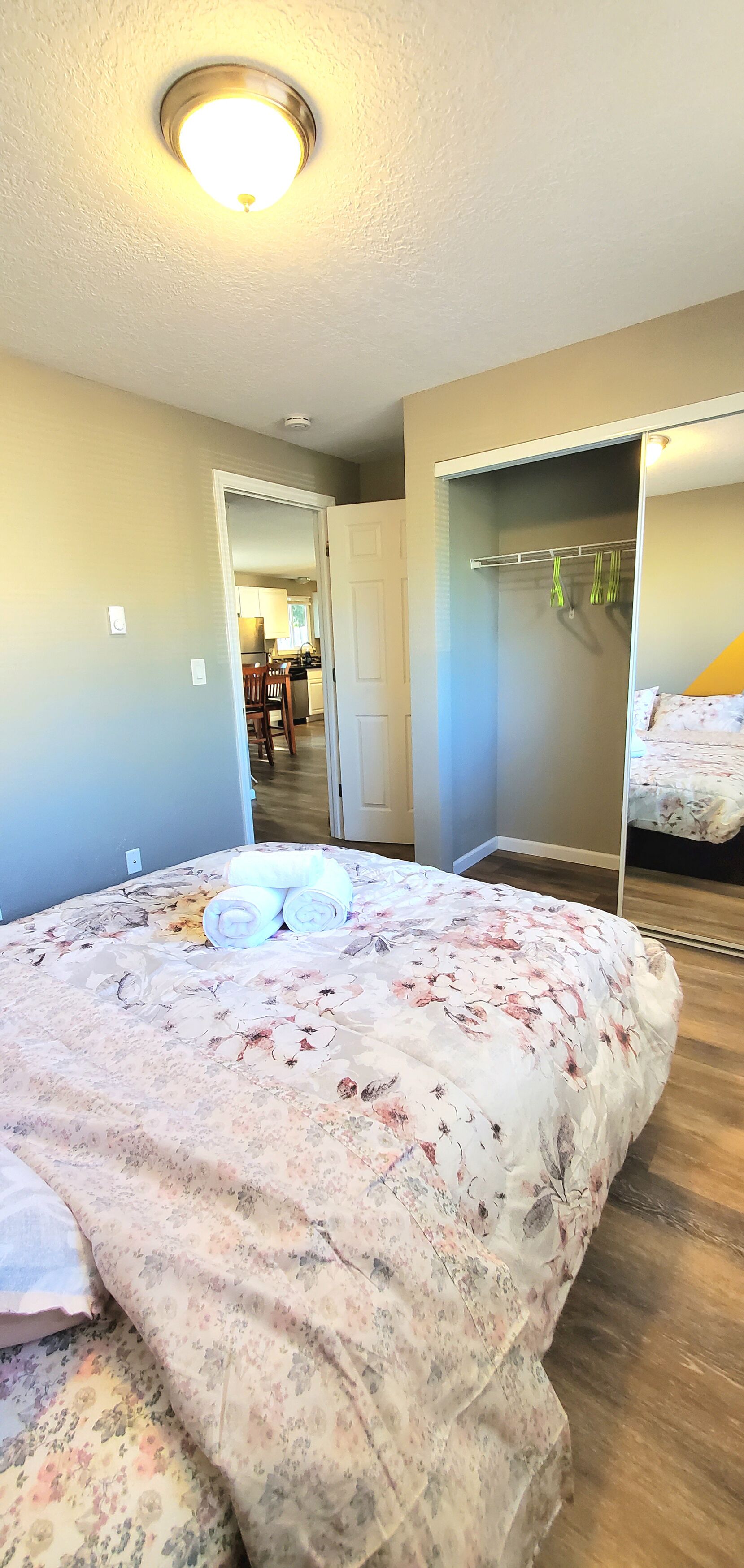 A bedroom with a floral bedspread, a closet with hanging clothes, and a door leading to a kitchen area.