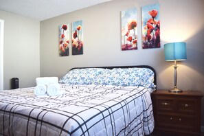 A neatly made bed with a floral-patterned bedspread and pillows, a bedside lamp, and three wall-mounted paintings of red flowers. - Duplex with Backyard and Putting Green (Beaverton)