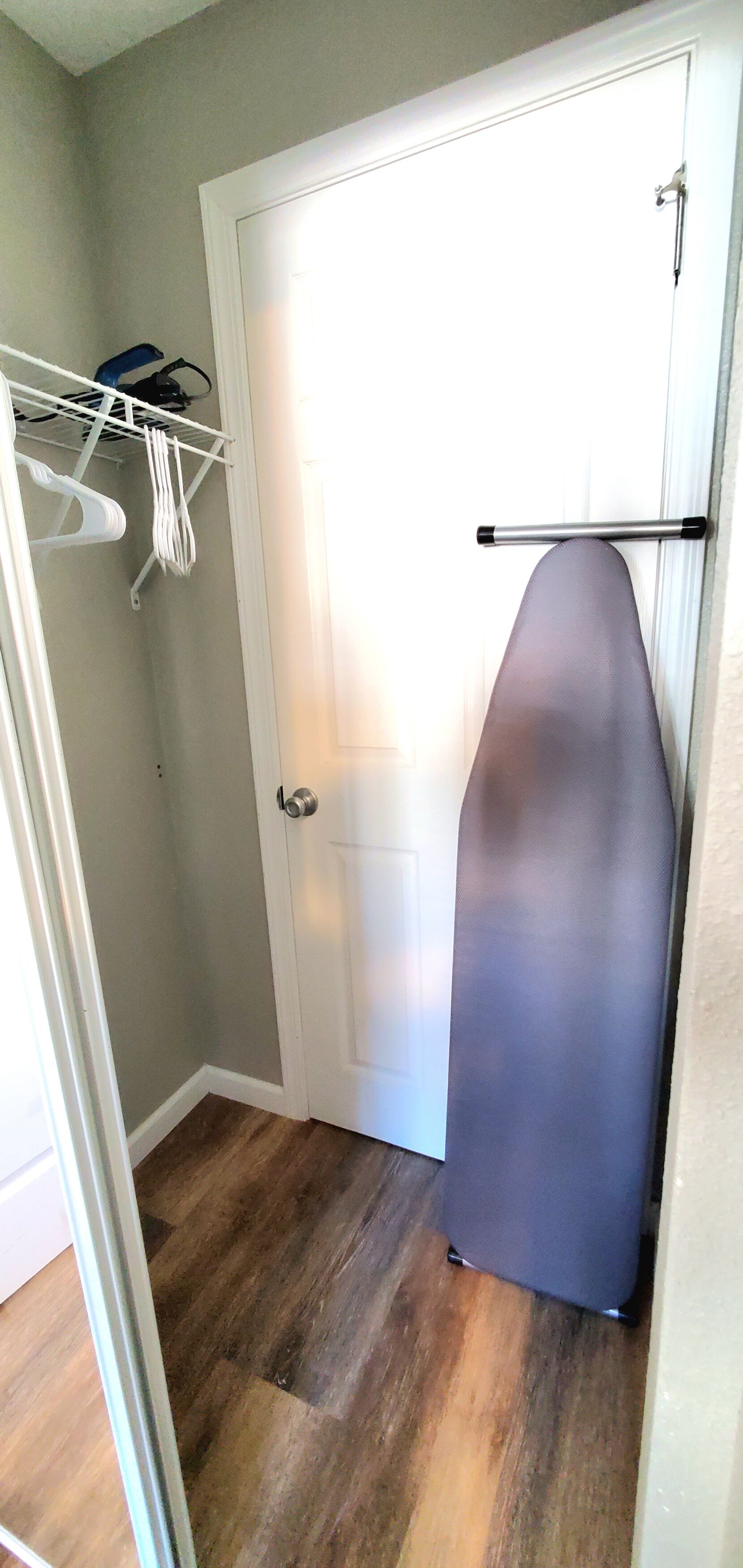 A narrow hallway with a white door, a hanging ironing board, and a white plastic shelf with items.