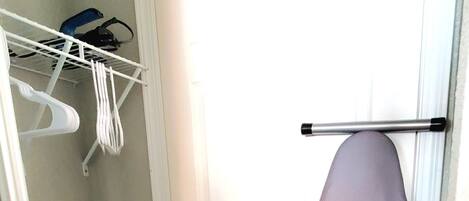 A narrow hallway with a white door, a hanging ironing board, and a white plastic shelf with items.