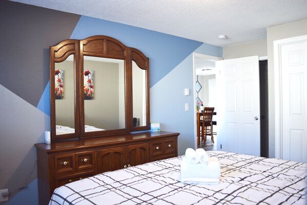 Une chambre à coucher avec un grand lit, une commode en bois, un miroir et une porte donnant sur une autre pièce.