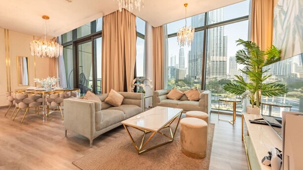 A spacious living room with a large window, a dining area, and a modern chandelier.