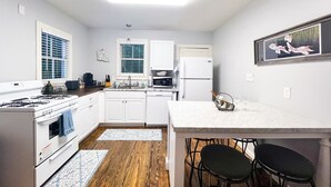 A compact kitchen with white cabinets, a stove, and a refrigerator. There are two bar stools and a small dining table.