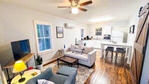 A living room with a sofa, a coffee table, a TV, and a lamp. There is a kitchen area with a stove, sink, and bar stools.