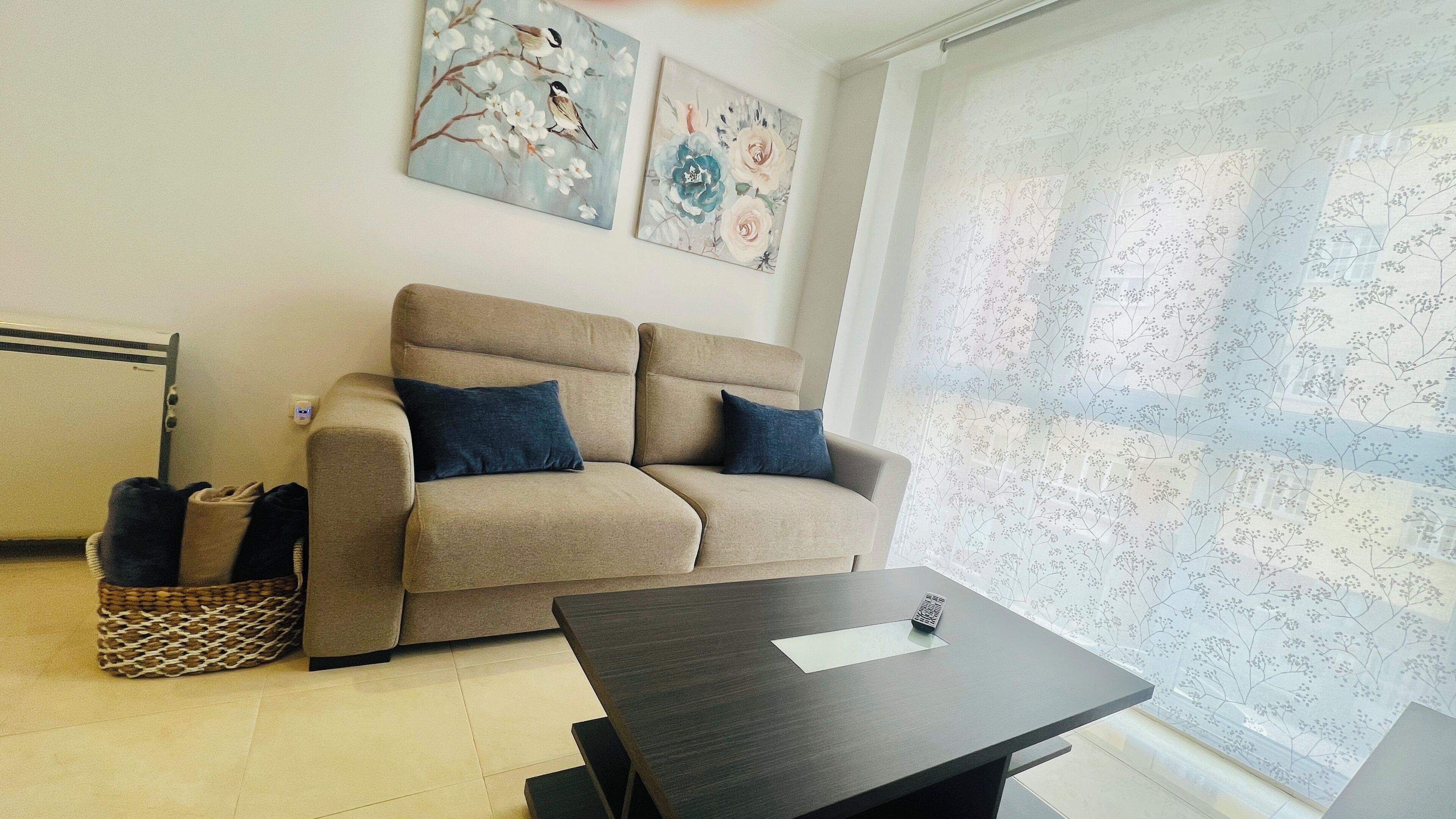 A living room with a beige sofa, two dark blue cushions, a small table, a basket, and two floral paintings on the wall.
