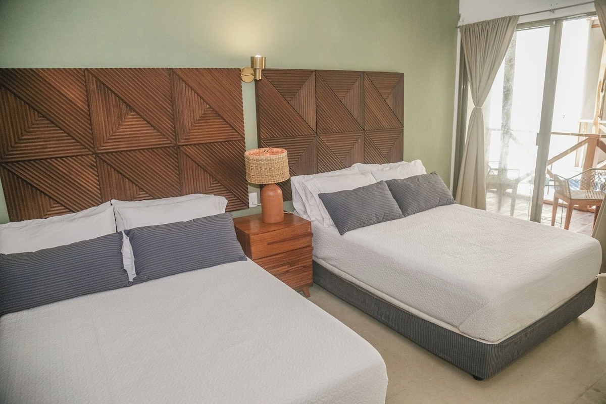 A hotel room with two beds, a wooden headboard, a nightstand with a lamp, and a view of the outdoors.