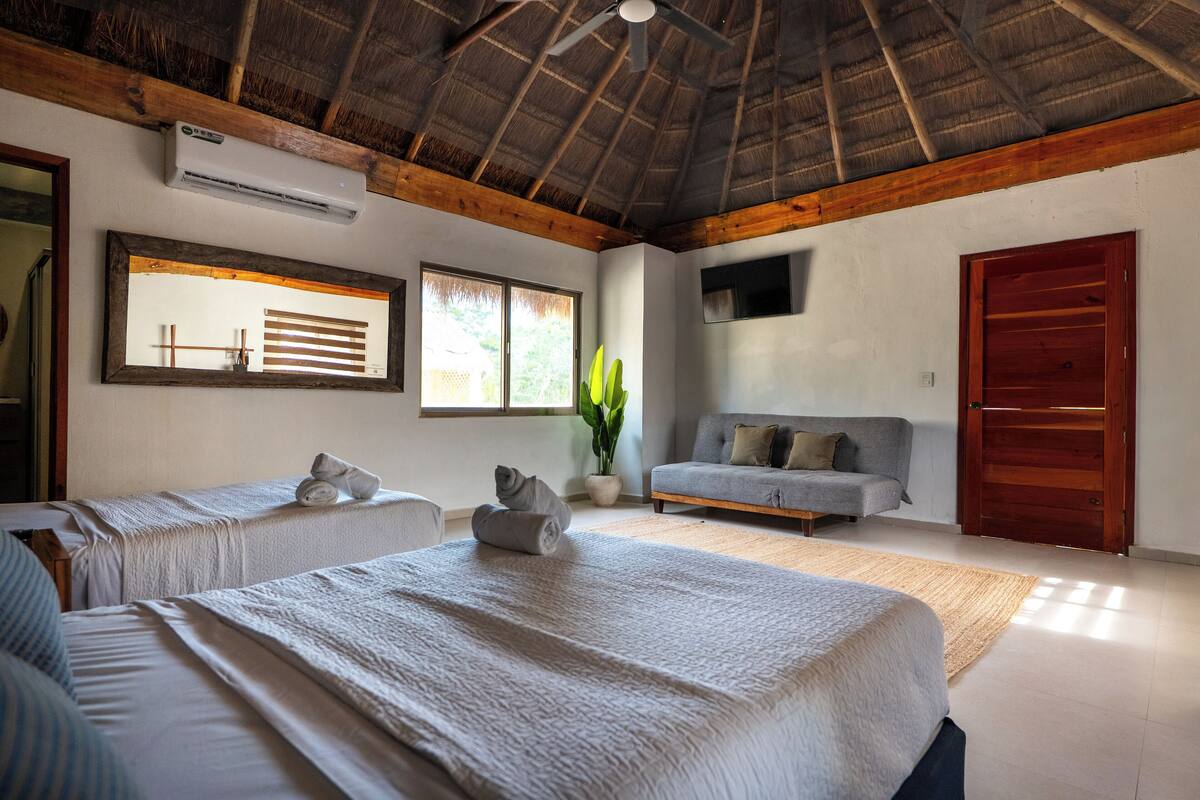 A hotel room with two beds, a sofa, a ceiling fan, a large window, and a door.