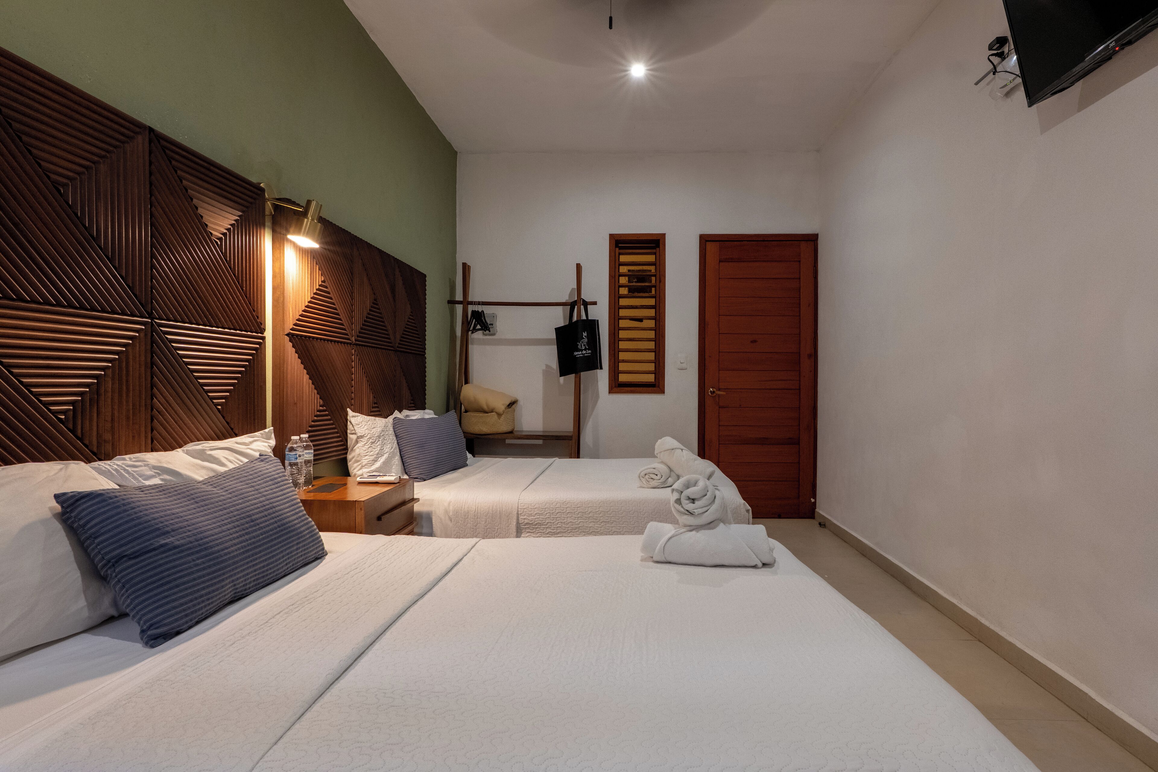 A hotel room with two beds, wooden headboards, a bedside table with a lamp, a wardrobe, and a television mounted on the wall.