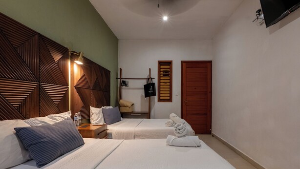 Habitación de hotel con dos camas, cabeceros de madera, una mesita de noche con lámpara, un armario y un televisor montado en la pared.