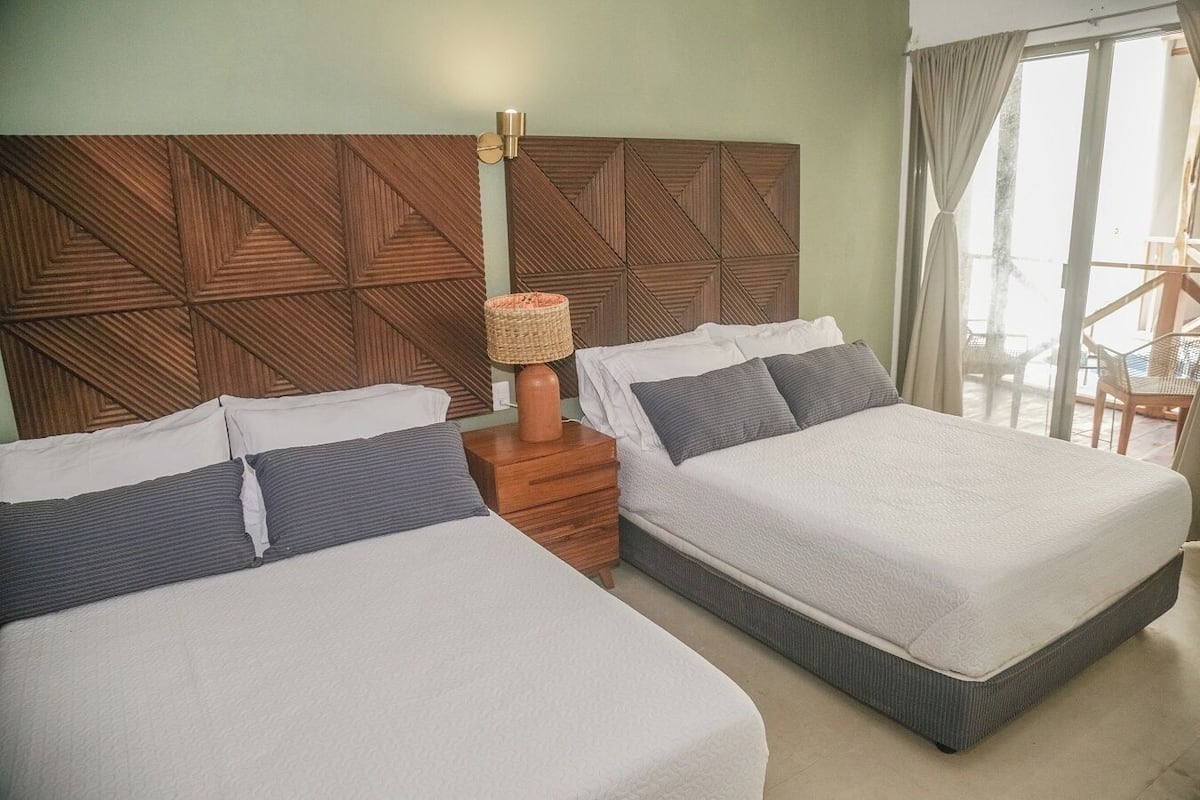 A hotel room with two beds, a wooden headboard, a nightstand with a lamp, and a view of the outdoors.