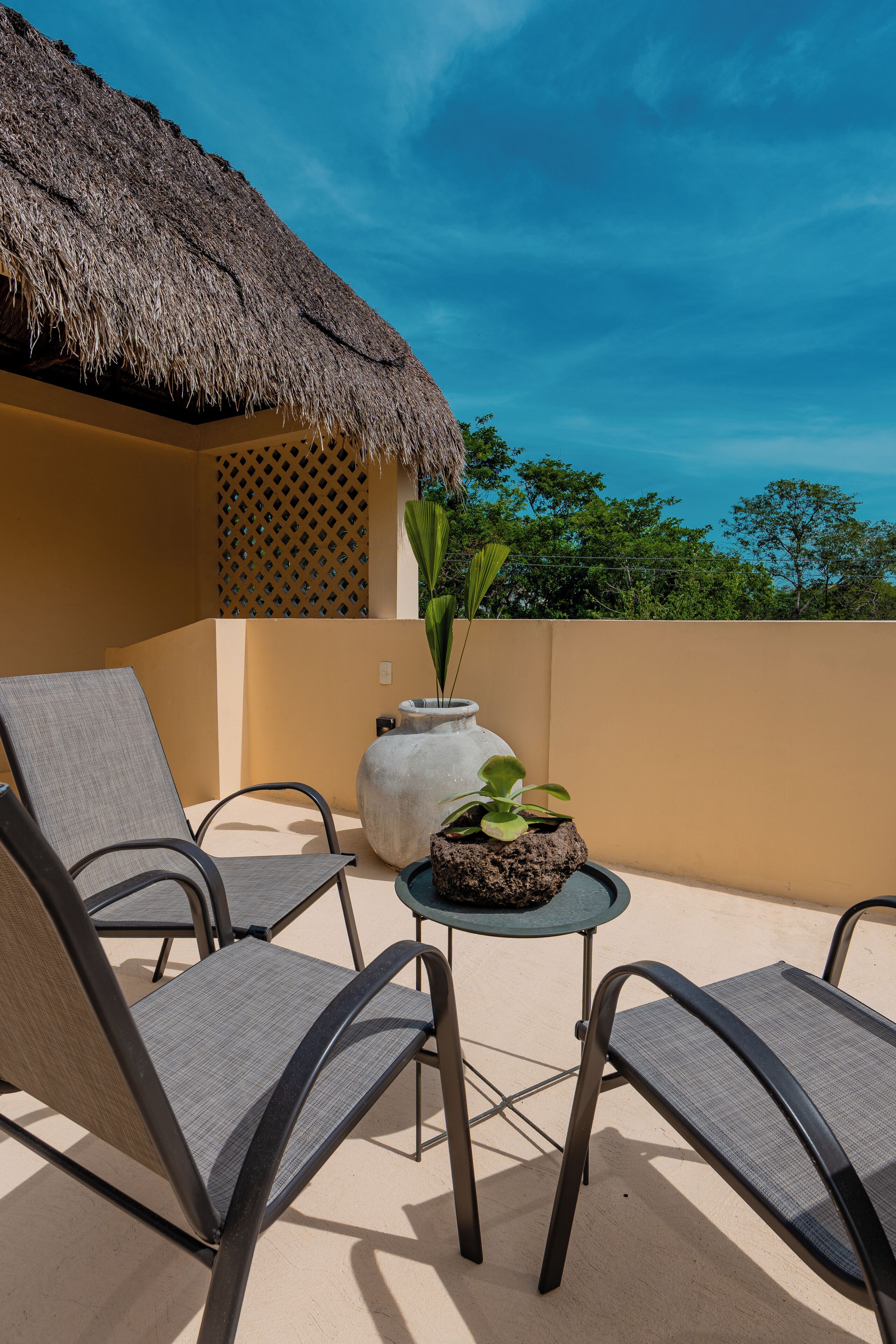 A balcony with two chairs, a small table, a potted plant, and a thatched roof structure.
