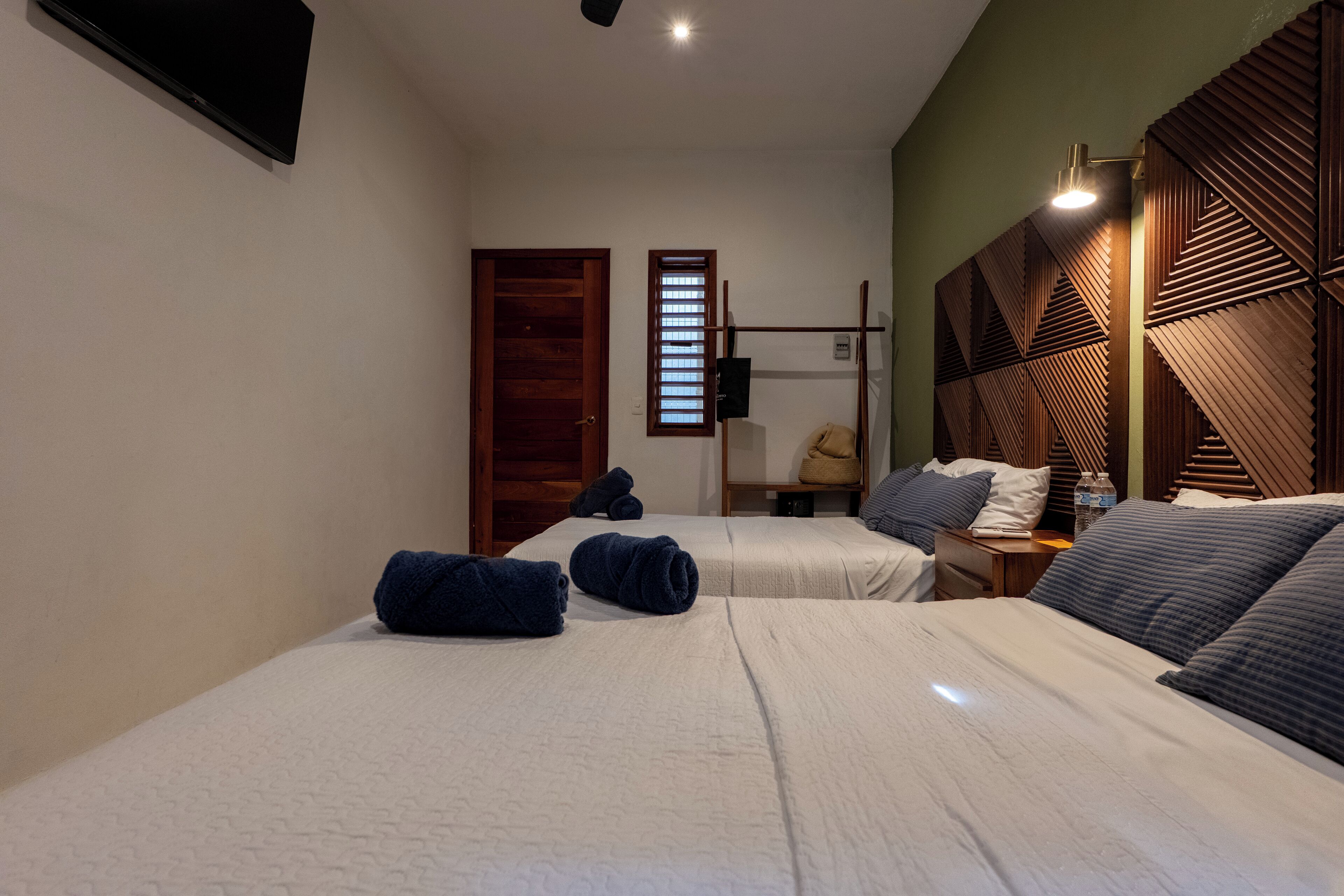 A hotel room with two beds, a wooden headboard, a nightstand, a lamp, and a window with blinds.