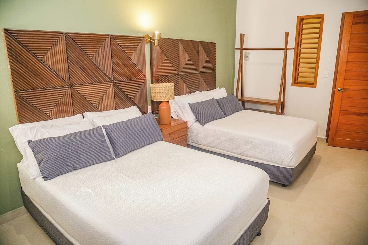 A room with two beds, a wooden headboard, a nightstand with a lamp, and a wooden door.