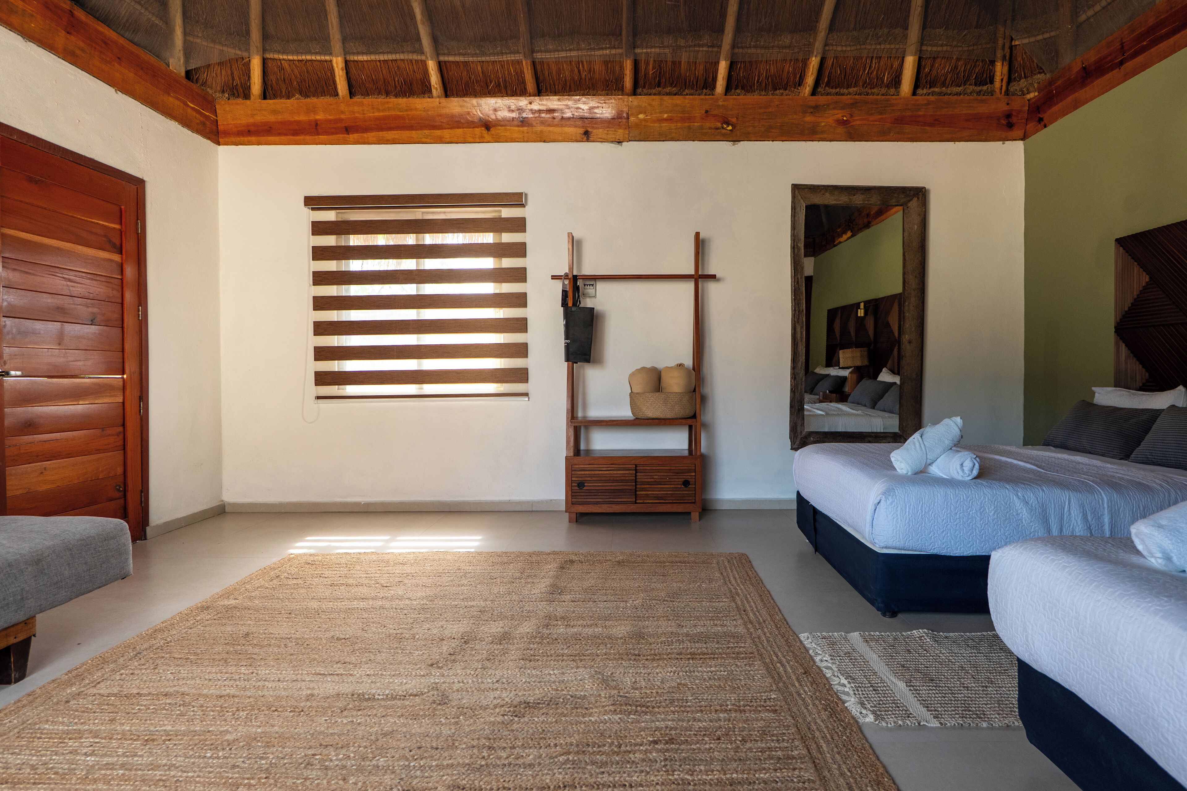 A room with two beds, a wooden bench, a woven rug, a wooden shelf with baskets, and a large mirror.