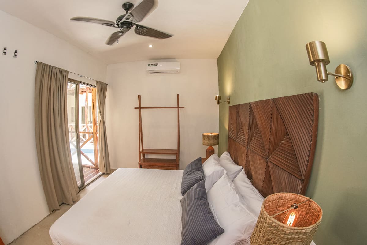 A bedroom with a bed, a ceiling fan, an air conditioning unit, a nightstand with a lamp, and a decorative wall panel.