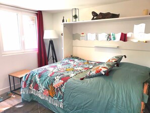 Une chambre à coucher comprenant un lit, une table de chevet, une lampe, une fenêtre avec des rideaux et une étagère ornée d’objets décoratifs.