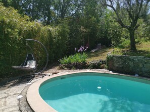 Une piscine entourée d’une végétation luxuriante, un fauteuil suspendu et un mur de pierre.