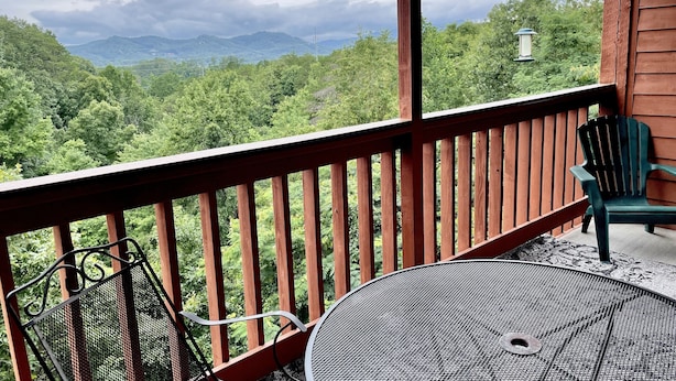Une terrasse en bois avec une table et des chaises en métal, offrant une vue sur un paysage verdoyant et luxuriant, avec des montagnes à l’horizon.