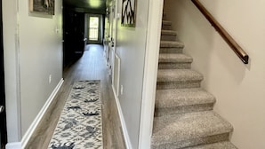 Un couloir avec un tapis à motifs, un escalier doté d’une rampe en bois et des tableaux encadrés accrochés aux murs.