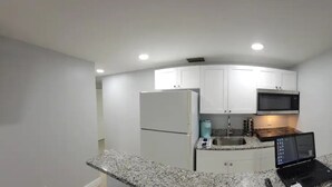 Cocina compacta con armarios blancos, microondas integrado, pileta de acero inoxidable y una computadora portátil sobre la encimera.