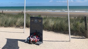 Deux drapeaux flottant sur des mâts, une plaque commémorative et une vue sur la plage.