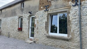 Un bâtiment en pierre dont les murs présentent un mélange de parties intactes et de parties endommagées, avec une porte bien visible et une fenêtre agrémentée d’une jardinière.