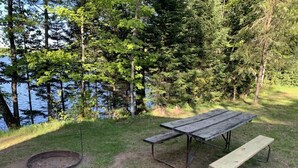 A picnic table with benches, a fire pit, and a grassy area near a lake surrounded by trees. - Gresham Shores - Woodies Perch - PET FRIENDLY (Arbor Vitae)