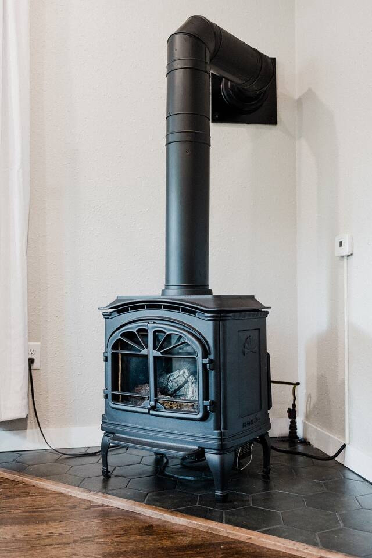 Een zwarte houtkachel met schoorsteen, voor een witte muur met betegelde vloer.