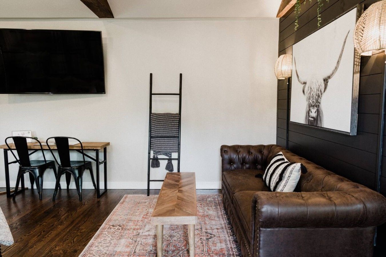 Een woonkamer met een bruine leren bank, een houten salontafel, een aan de muur bevestigde televisie en een vloerkleed met een patroon.