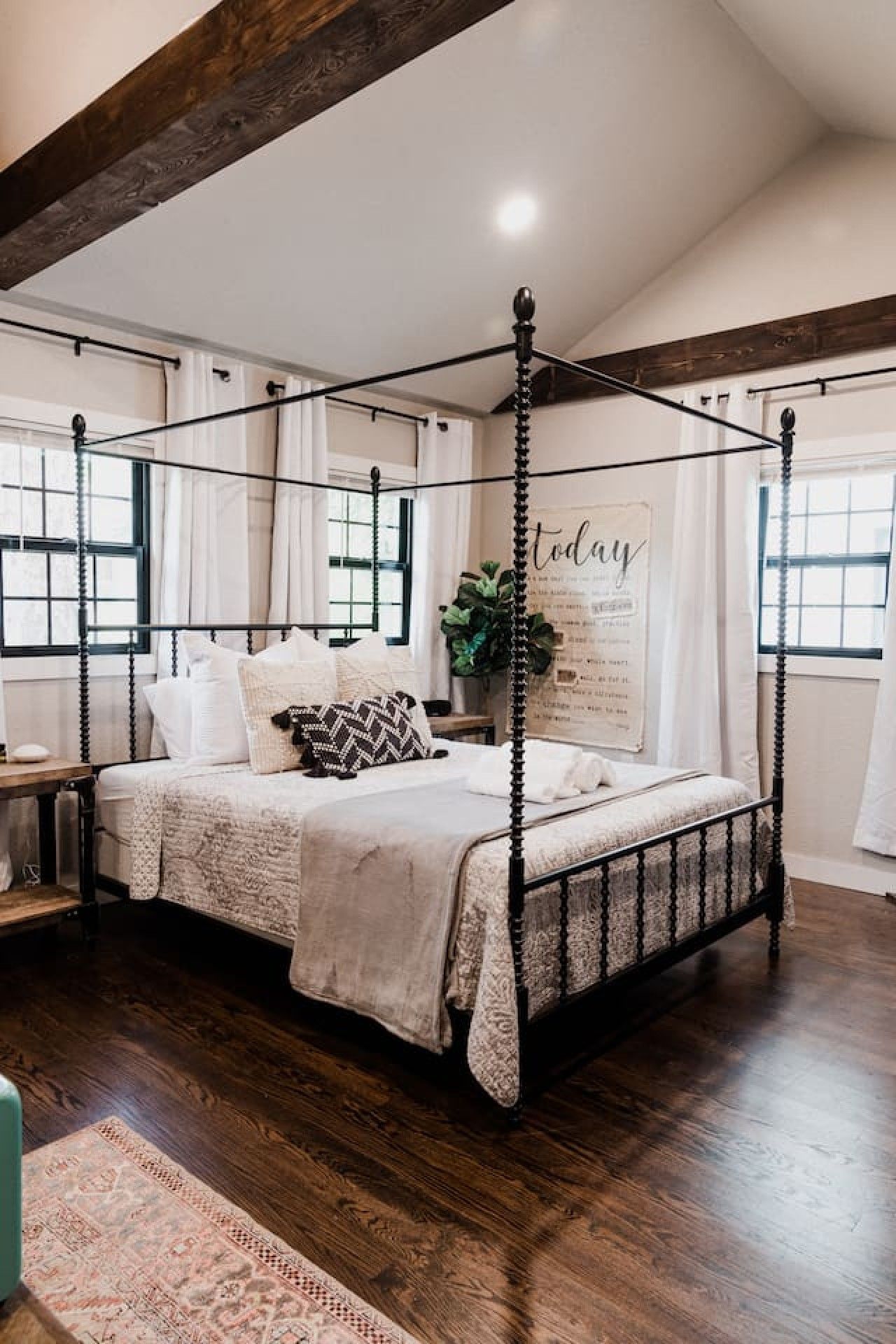 A four-poster bed with a canopy, a bedside table, a potted plant, and a wall with a poster.