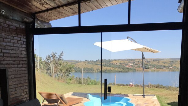 A modern room with a large sliding glass door leading to a pool area with a view of a lake and hills. - Chalé Mirante Reservations only through the website ChalesTemporada.com (Alexânia)