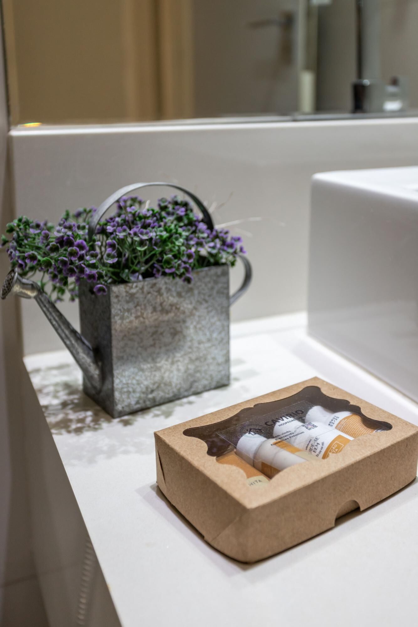 Une salle de bain avec un petit arrosoir en métal contenant des fleurs violettes, une boîte en carton avec un produit, et un comptoir blanc.