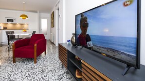 Un salon moderne avec un grand téléviseur à écran plat, un fauteuil rouge, un meuble de rangement en bois et une cuisine en arrière-plan.
