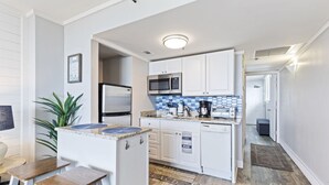 A compact kitchen with white cabinets, a microwave, a refrigerator, and a coffee maker. There is a small dining area with stools and a plant.