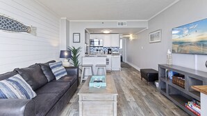 A modern living room with a dark leather sofa, a coffee table, and a TV stand with books. A kitchen is visible in the background with white cabinets and a microwave.