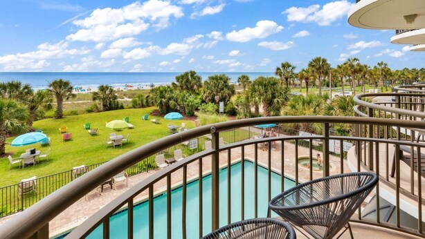 A pool area with a view of the beach, outdoor seating, and a poolside railing.