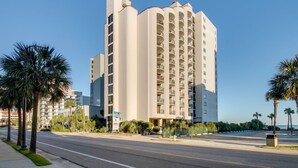 A tall, beige multi-story hotel with balconies, located near a palm tree-lined street.