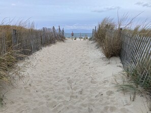 Ein sandiger Küstenpfad, gesäumt von Holzzäunen und hohem Gras, der zum Meer führt.