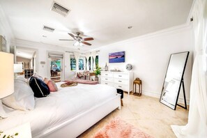 A spacious bedroom with a large bed, a ceiling fan, a dresser, a mirror, and a view of the outdoors.