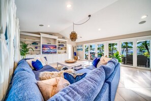 A spacious living room with a large blue sofa, a coffee table, and a TV on the wall. There is a balcony with a view of palm trees and a pool.