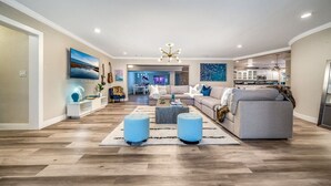 A spacious living room with a large sofa, two ottomans, a coffee table, and a TV mounted on the wall. - NEW! 5 BED/5 MIN TO BEACH/HTD POOL/HOT TUB/BAR (Oakland Park)